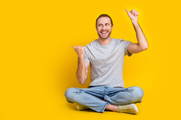 Full size photo of positive man gray t-shirt pants sitting on floor look indicating empty space sale isolated on yellow color background