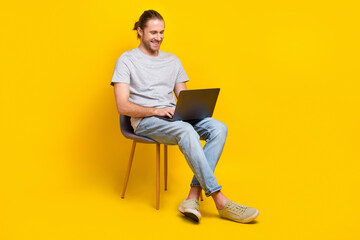 Full size photo of handsome clever man gray t-shirt pants sitting on chair writing code distance work isolated on yellow color background