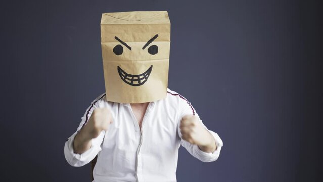 A man in a white shirt with a bag on his head, with an angry smiley face drawn, gloats emotionally while sitting in a white chair. Laughs, showing fists. Emotions and gestures.