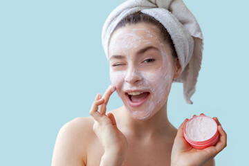 Beauty portrait of young woman in towel on head applying white nourishing mask or creme on face...