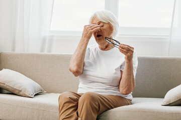 Elderly woman severe eye pain sitting on the couch, health problems in old age, poor quality of...