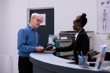 Obraz premium Senior patient standing at hospital reception counter paying medical examination with smartphone using contactless, talking with receptionist in lobby. Old man making payment after having consultation