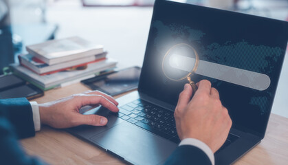 search engine concept, businessman working on computer laptop holding magnifying glass and search icon on busy table in office