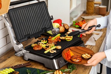 Woman cooking tasty vegetables on modern electric grill at table in kitchen