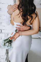 The groom hugs the brunette bride with curly hair, holding a bouquet of flowers behind her back, standing indoors. Wedding photography, portrait.