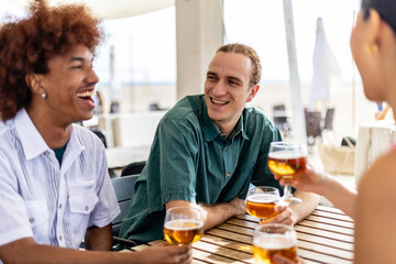 Young group of multiracial people drinking cold beer at beach bar terrace on vacation. Millennial diverse friends hanging out enjoying summer day at beach party. Summertime and friendship concept