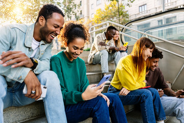 Group of multiracial young people using mobile phone devices sitting outdoors. Millennial happy...