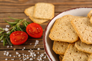 Bowl with tasty crackers, salt, tomatoes and rosemary on wooden background
