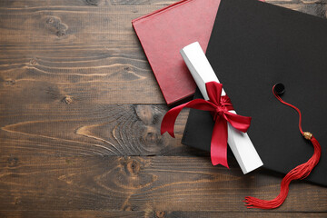 Diploma with red ribbon, graduation hat and book on wooden table