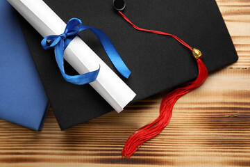 Diploma with blue ribbon, graduation hat and book on wooden table