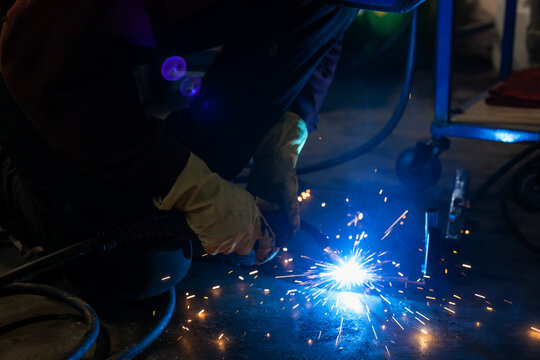 A hardworking mechanic in a welding mask and oil-stained clothing is welding parts in an auto repair shop or car workshop. Blue flashes of light and bright orange sparks are flying in all directions.