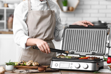 Woman cooking tasty vegetables on modern electric grill at table in kitchen, closeup
