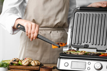 Woman cooking tasty vegetables on modern electric grill at table in kitchen, closeup