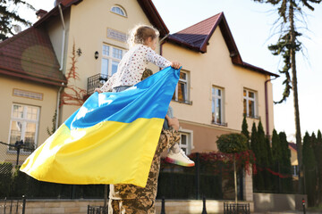 Soldier in military uniform carrying his daughter on shoulders and Ukrainian flag on city street,...