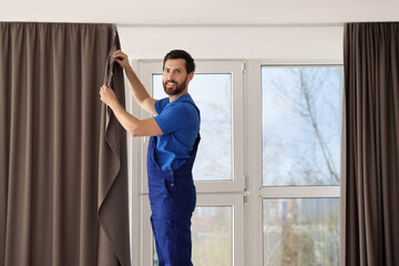 Worker in uniform hanging window curtain indoors