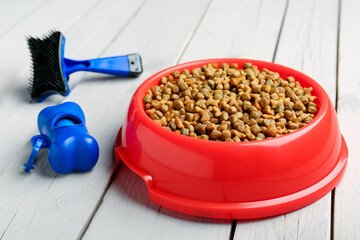 Bowl of dry pet food, waste bags and grooming brush on light wooden background