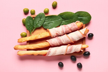 Wooden board of tasty Italian Grissini with bacon on pink background