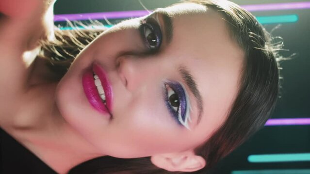 Vertical video. Disco makeup. Fashion girl. Retro 90s beauty. Confident teen model face with blue color eyeshadow pink lips artistic look posing in neon light on black.