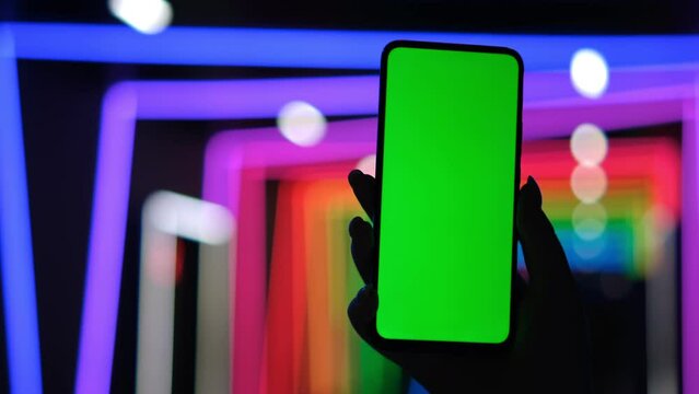 African American woman holds smartphone in her hand and touches green screen chroma key. Close up of a phone in a horizontal position against background of bright multicolored neon lights. Slow motion