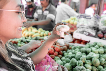 A young Hispanic woman is smiling while looking at a mushroom in a street market
