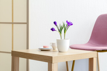 Pot with beautiful crocus flowers, cup and tasty macaroon on table near white wall