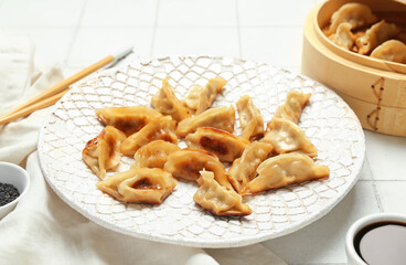 Plate with tasty Chinese jiaozi, sesame and bamboo steamer on white tile background