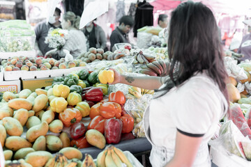 Close up of a young Hispanic woman is holding a yellow pepper while doing grocery shopping in a street market