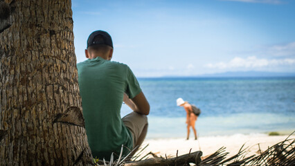 a man, on the beach, looks at the silhouette of a woman by the water, a tropical island