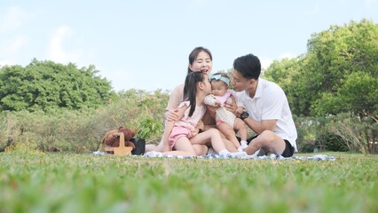 Cute asian girl kissing her little sister's cheek while sitting picnic with parents in the park.