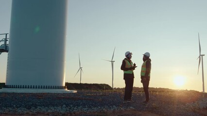 Wide shot of two renewable energy engineers in uniform and helmets standing with tablet at wind farm discussing turbines in morning, sunrise in background. Sustainable future, green energy concepts