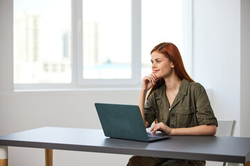 red-haired woman smiling thoughtfully sitting at work at a laptop