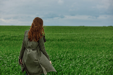 a red-haired woman in a long raincoat stands in a green field in cloudy weather in a strong wind