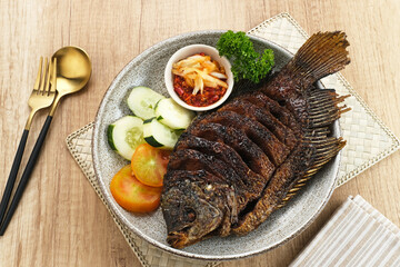 Gurame Bakar Madu, Grilled Gourami with honey and soy sauce. Served with sambal (chilli sauce), cucumber and tomatoes in white plate.   
