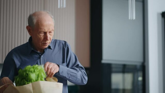 Elderly Man Putting Out Bread And Vegetables From Bag After Visit Of Supermarket In Weekend Portrait