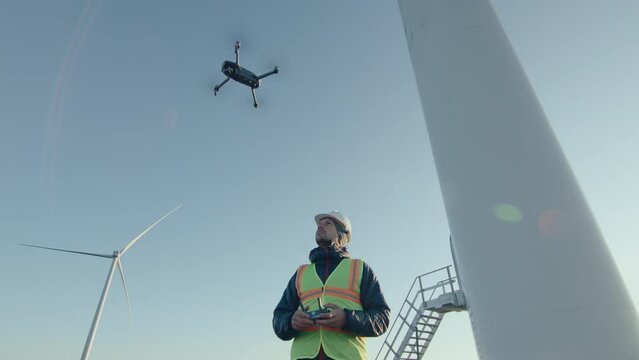Engineer in workwear and hardhat launching a drone from his hand and using controller, inspecting wind turbine at the wind power plant. Low angle shot