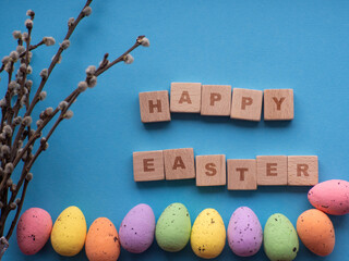 Happy Easter concept. Easter decorations on blue background with copy space. Easter greating card. Flat lay. Colorful eggs and Happy Easter inscription on wooden cubes. Celebration of spring holidays.