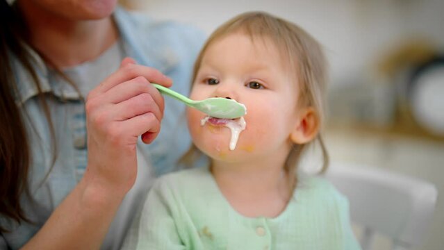 Mom feeding her baby with milk porridge at home. Portrait of baby toddler sitting in mother's hands. Raising child, motherhood, parenthood love and care in family, child's nutrition food concept.