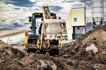 Obraz premium A universal excavator is working on a construction site against a cloudy sky. Earthworks at a construction site. Modern earthmoving equipment. The contractor is doing construction work.