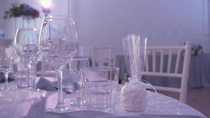 Empty glasses, napkins and gift wrapped craft marshmallows stand on table in beautiful blue room. Nearby are chiavari chairs, spotlight flickers beautifully on counter next to flower arrangement. - Powered by Adobe