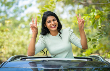 Happy smiling woman showing victory sign by looking at camera on car sunroof - concept of...
