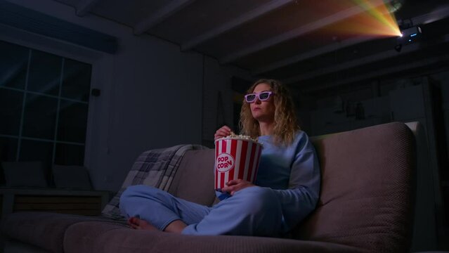Young woman enjoying a movie with popcorn. Caucasian woman watching 3D movie in her home theater, with multicolored projector beams in the background.. High quality 4k footage