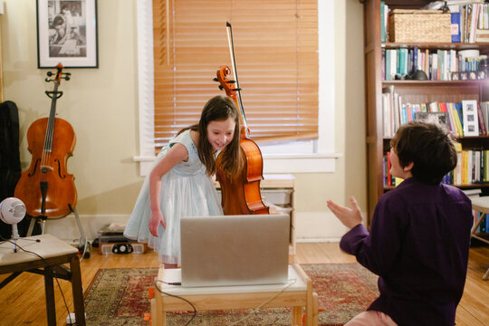 A little girl with cello bows in front of computer while brother claps