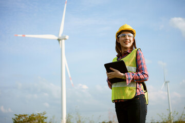 Asian woman in yellow helmet working with digital tablet at renewable energy farm. Female inspector controlling functioning of wind turbines outdoors.