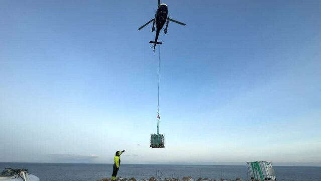 Medium shot of a helicopter bringing in supplies on a longline near the ocean with an operator directing and ready to receive