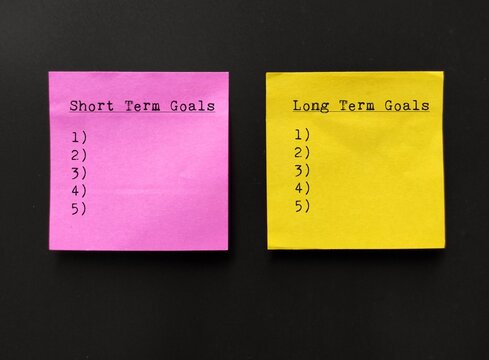 Pink and yellow note stick on wall with lists of SHORT TERM GOALS and LONG TERM GOALS, concept of goals setting goals, easy ones to achieve soon and bigger ones to accomplish in the future