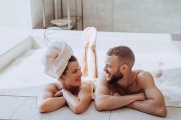 Happy caucasian couple enjoying holidays at spa laying in jacuzzi at hotel, vacation. Pretty woman in love looks at  bearded boyfriend, smiles. Hispanic guy toothy smiling enjoying weekend with wife. - obrazy, fototapety, plakaty