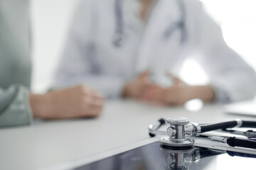 Doctor and patient are sitting and discussing something at the desk in the clinic office. The focus is on the stethoscope lying on the table, close up. Perfect medical service and medicine concept.