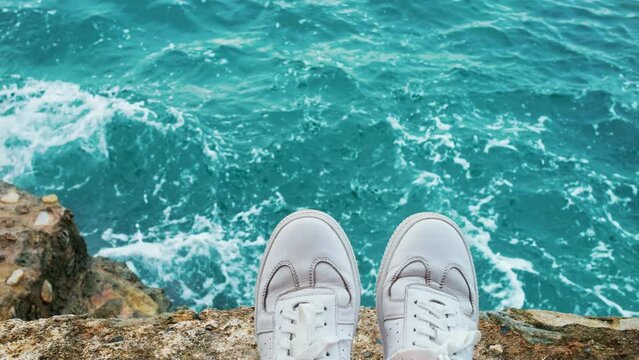 Traveler or adventurer, nomad urban explorer sits on cliff or rock in white sneakers, watches waves crush and roll over shore. Influencer or blogger lifestyle. Feet hanging from cliff waves on coast