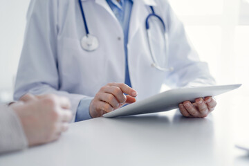 Doctor and patient sitting at the table in clinic. The focus is on female physician's hands using tablet computer, close up. Medicine and healthcare concept