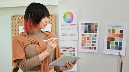 Young asian woman designer illustrator standing in modern design studio and using digital tablet - obrazy, fototapety, plakaty